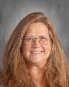 Smiling woman with long, wavy brown hair and glasses, set against a soft gray background. The image conveys a friendly and approachable demeanor, suitable for a professional profile or educational context.