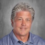Professional headshot of a middle-aged man with gray hair, wearing a blue checkered shirt, smiling against a textured gray background. Ideal for educational or professional profiles.