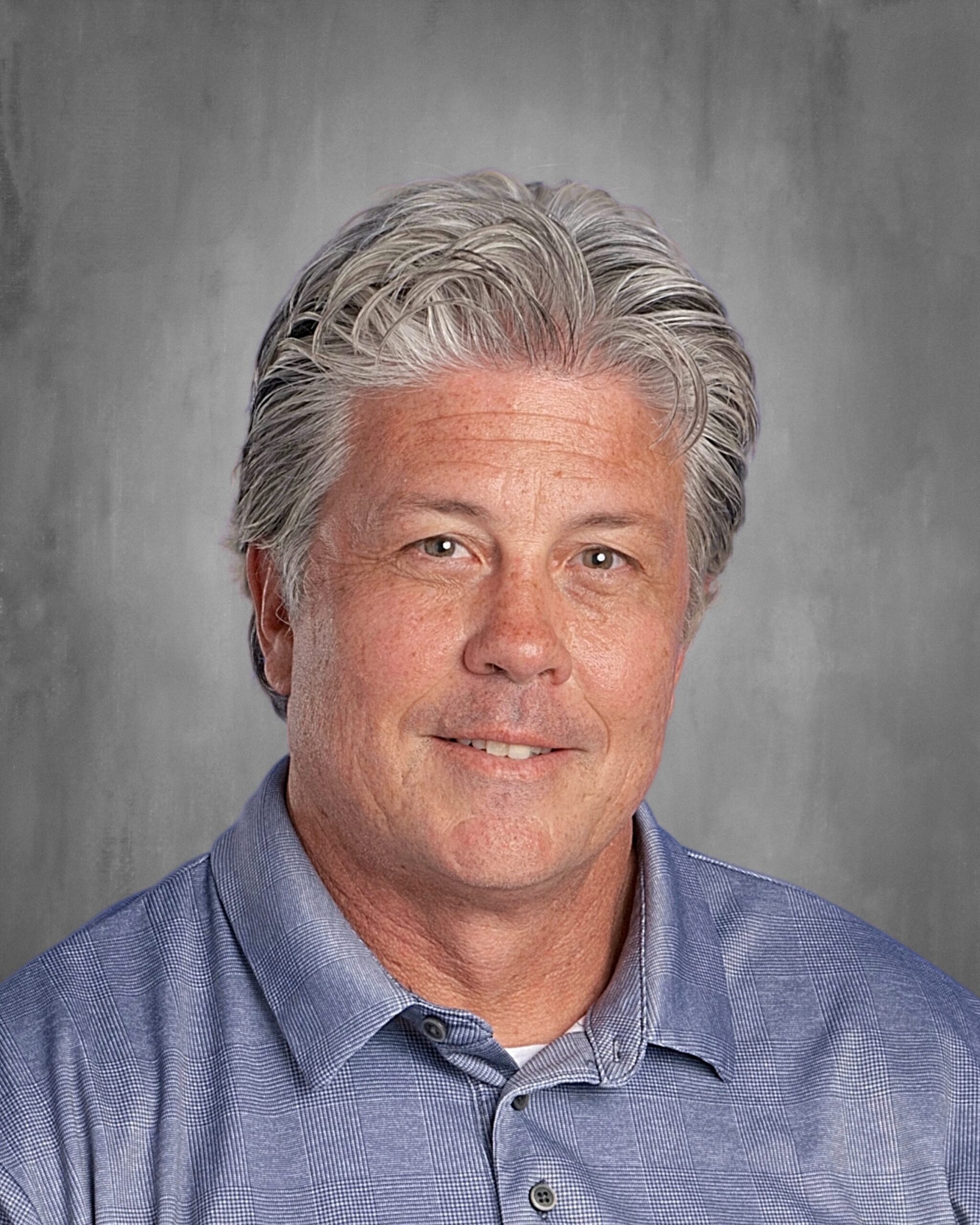 Professional headshot of a middle-aged man with gray hair, wearing a blue checkered shirt, smiling against a textured gray background. Ideal for educational or professional profiles.