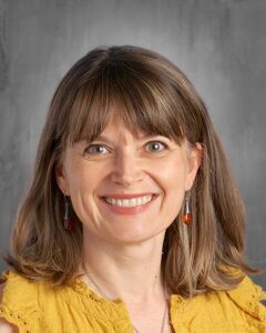 Professional headshot of a smiling woman with medium-length brown hair and earrings, wearing a yellow blouse. The background is a soft gray, enhancing her friendly expression.