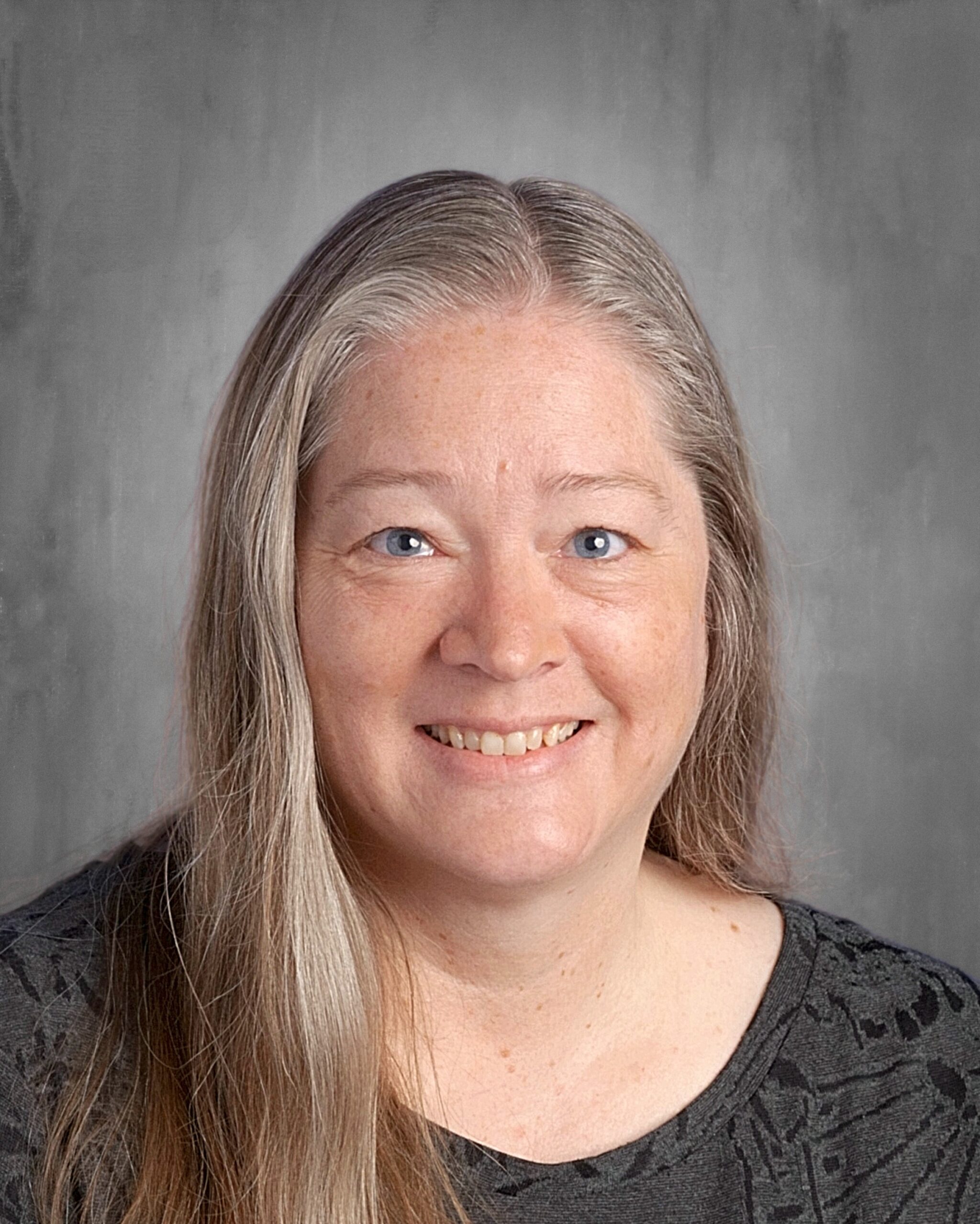 Portrait of a smiling woman with long gray hair, wearing a dark patterned shirt, against a neutral gray background. Ideal for showcasing a friendly and approachable personality.