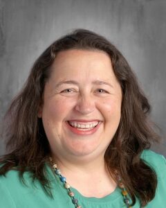 Smiling woman with long dark hair wearing a green blouse and a colorful beaded necklace, set against a neutral background.