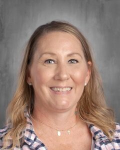 Professional headshot of a smiling woman with long, wavy blonde hair, wearing a plaid shirt and gold jewelry, set against a neutral gray background.