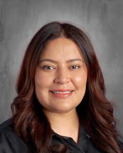Professional headshot of a woman with long, wavy dark hair, smiling against a neutral gray background, wearing a black top. Ideal for business profiles or educational contexts.