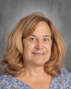 Professional headshot of a smiling woman with wavy, shoulder-length hair, wearing a blue floral blouse, set against a neutral gray background. Ideal for showcasing a teacher or educator's profile.