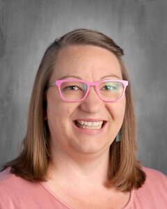 Smiling woman with shoulder-length hair wearing pink glasses and earrings, posing against a gray background.