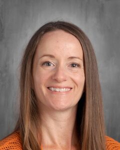 Professional headshot of a woman with long brown hair, smiling and wearing an orange top, set against a neutral gray background.