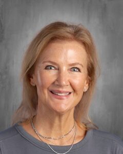 Smiling woman with blonde hair wearing a gray shirt and multiple silver necklaces, set against a textured gray background. Suitable for use in professional or educational contexts.