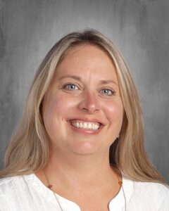 Smiling woman with long blonde hair and blue eyes, wearing a white blouse, against a gray background. Ideal for educational or professional profile.