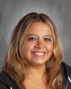 Smiling woman with long, wavy hair wearing a black hoodie, set against a gray background.