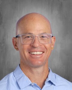 Smiling man with glasses and earrings wearing a light blue shirt, against a gray background.