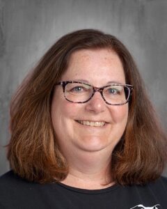 Portrait of a middle-aged woman with shoulder-length brown hair and glasses, smiling against a neutral background. She is wearing a black shirt, conveying a friendly and approachable demeanor.