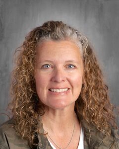 Portrait of a smiling woman with curly hair, wearing a camouflage jacket, against a neutral gray background.