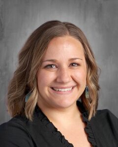 Smiling woman with shoulder-length wavy hair, wearing a black top and colorful earrings, against a gray background.