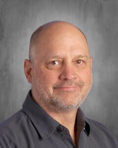Headshot of a middle-aged man with short, bald hair and a light beard, wearing a dark button-up shirt, smiling against a grey background.