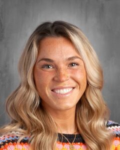 Smiling woman with blonde, wavy hair and freckles, wearing a colorful knitted sweater, against a neutral gray background.