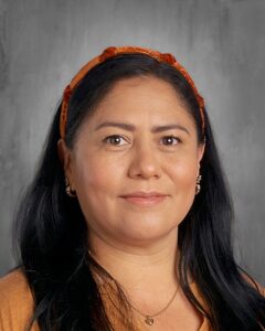 Portrait of a woman with long black hair wearing a brown top and an orange headband, set against a gray background. She has subtle makeup and is adorned with gold earrings and a necklace.