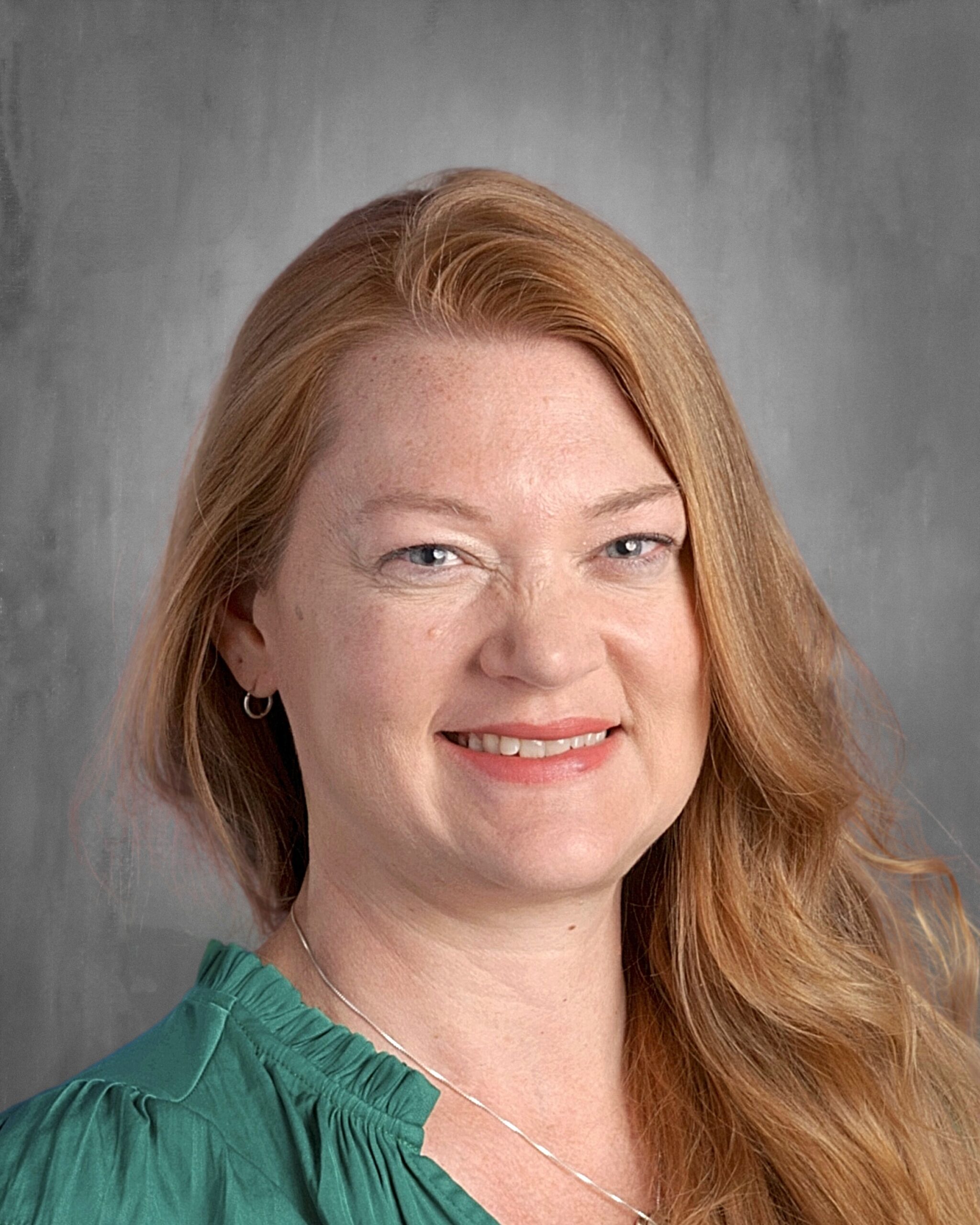 Portrait of a woman with long, wavy red hair and blue eyes, wearing a green blouse, smiling against a neutral gray background.