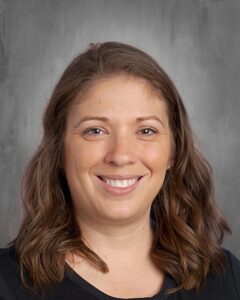 Portrait of a smiling woman with wavy brown hair, wearing a black shirt, against a textured gray background. Ideal for educational or professional profiles.