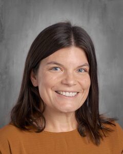 Smiling woman with long brown hair wearing a brown shirt against a gray background, showcasing a friendly and approachable demeanor.