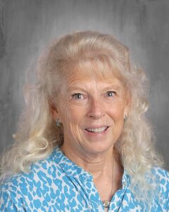 Portrait of a smiling woman with curly blonde hair, wearing a blue and white patterned blouse, against a gray background.