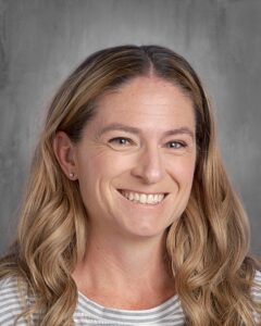 Smiling female teacher with long, wavy hair wearing a striped shirt, set against a neutral gray background.