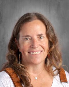 Smiling woman with long, wavy hair wearing earrings and a white blouse with brown overalls, posing against a gray background.