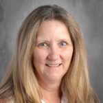 Smiling woman with long, light brown hair and blue eyes, wearing a white blouse, against a neutral gray background. Suitable for professional or educational contexts.