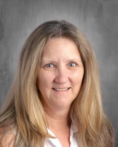 Smiling woman with long, light brown hair and blue eyes, wearing a white blouse, against a neutral gray background. Suitable for professional or educational contexts.