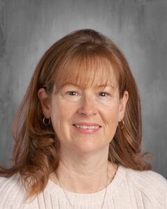 Professional headshot of a woman with shoulder-length reddish-brown hair, wearing a light sweater, smiling against a neutral gray background. Ideal for educational or professional profiles.