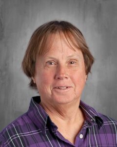 Portrait of a middle-aged woman with short brown hair, wearing a purple plaid shirt, smiling against a neutral gray background.