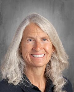 Smiling woman with long, wavy gray hair wearing a black shirt, set against a neutral gray background. Ideal for showcasing friendly and approachable professional images.