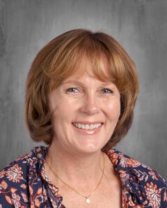 Professional headshot of a woman with shoulder-length, wavy brown hair, wearing a floral blouse. She is smiling and appears friendly, set against a neutral gray background. Ideal for educational or business profiles.