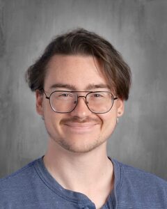 Smiling young man with glasses and medium-length hair, wearing a blue shirt, against a gray background.