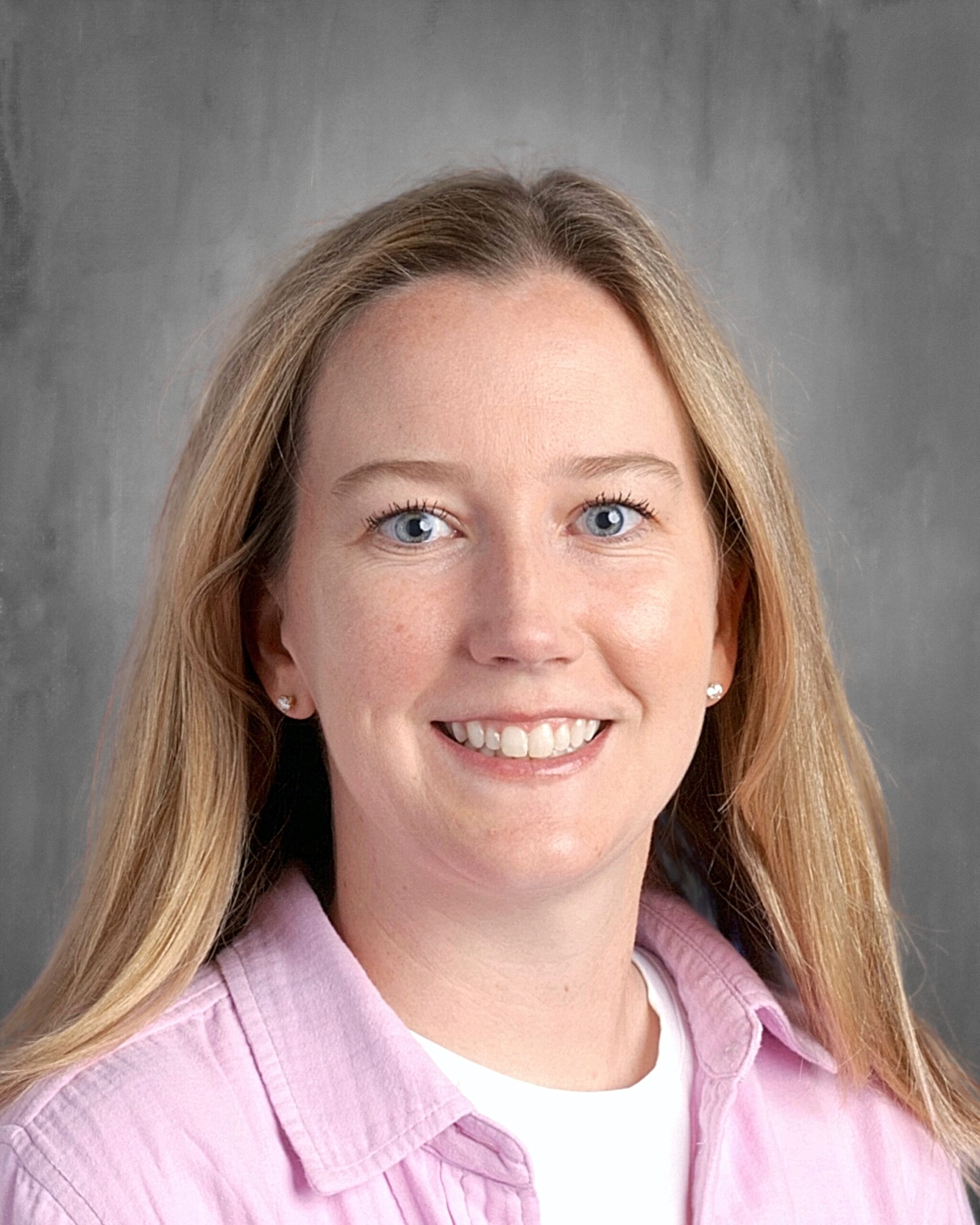 Smiling woman with long blonde hair and blue eyes, wearing a pink shirt over a white top, against a neutral background.