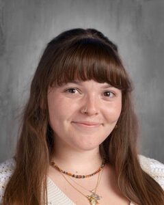 Portrait of a young woman with long brown hair and bangs, wearing a white sweater and layered necklaces, smiling against a neutral gray background.