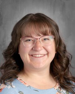 Smiling woman with curly hair wearing glasses and a floral blue shirt, posing against a neutral background.