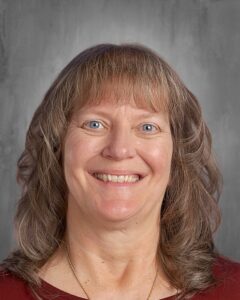 Smiling woman with long, wavy hair and blue eyes, wearing a maroon top and a necklace, against a gray background.