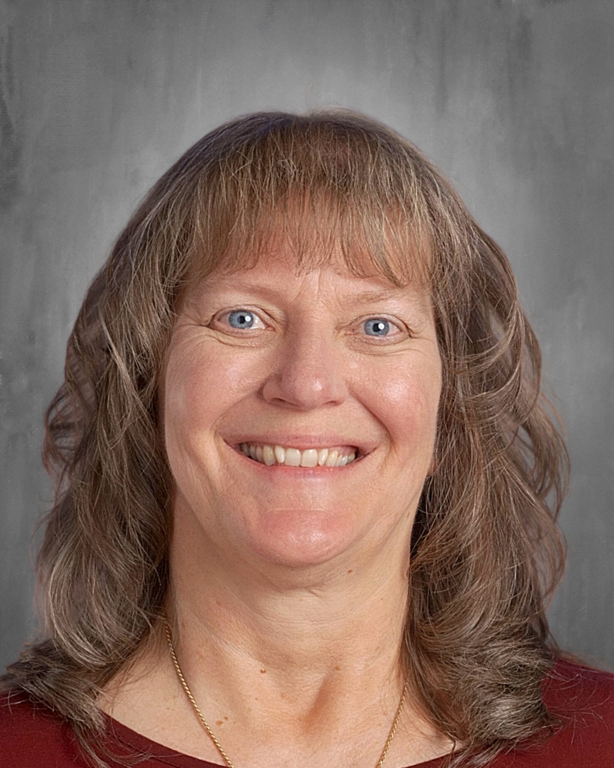 Smiling woman with long, wavy hair and blue eyes, wearing a maroon top and a necklace, against a gray background.