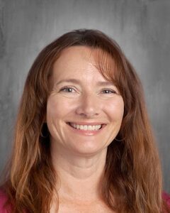 Portrait of a smiling woman with long, brown hair and a warm expression, set against a gray background. She is wearing a pink top and has hoop earrings. The image conveys a friendly and approachable demeanor, suitable for a professional profile or educational context.