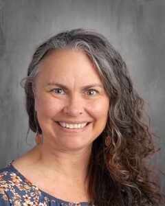 Smiling woman with long, curly gray hair wearing a floral blue blouse and dangling earrings, set against a neutral gray background.