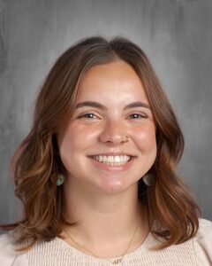 Smiling young woman with brown hair and earrings, wearing a light-colored top, against a gray background.