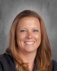 Professional headshot of a woman with long brown hair, smiling confidently against a neutral gray background. Ideal for use in business or educational contexts.