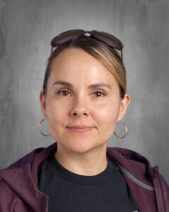 Portrait of a woman with light brown hair and hoop earrings, wearing a dark shirt and a purple jacket, with sunglasses resting on her head, set against a neutral gray background.