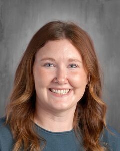 Portrait of a smiling woman with wavy red hair and a nose ring, wearing a teal shirt, set against a gray background. Ideal for showcasing a friendly and approachable demeanor.