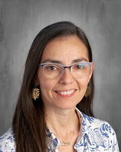 Professional headshot of a woman with long dark hair, wearing glasses and decorative earrings, smiling against a grey background. She is dressed in a blue and white patterned blouse and a gold necklace.