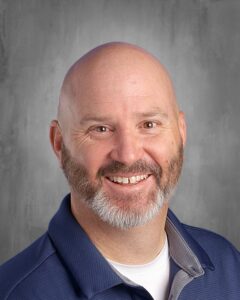 Headshot of a smiling man with a bald head and a gray beard, wearing a navy blue polo shirt against a neutral gray background.