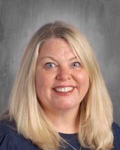 Smiling woman with long blonde hair and blue eyes, wearing a dark top, against a gray background.